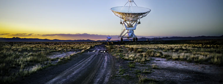 The steel lattice and mount of a large parabolic antenna against a dusk sky.